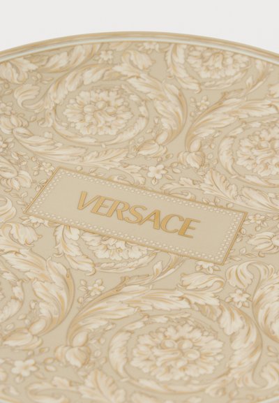 Μπεζ πιάτο Versace με περίτεχνα χρυσά λουλουδένια και φυλλώδη σχέδια και το "VERSACE" τυπωμένο στο κέντρο με χρυσό χρώμα.