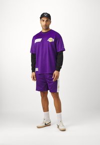 Maglia sportiva in rete viola con maniche lunghe nere, abbinata a pantaloni corti viola. Presenta loghi NBA e dettagli bianchi. Sneakers e calzini bianchi.