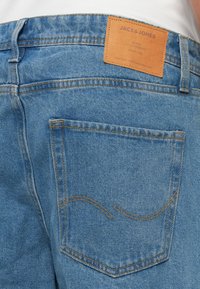 Nærbilde av en baklomme på blå denimjeans med oransje søm og en brun lær JACK & JONES merkeetikett over.