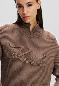 Brauner, gerippter Strickpullover mit hohem Kragen und gesticktem "Karl"-Design auf der Vorderseite. Die Textur wirkt dick und gemütlich.