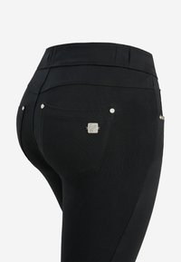 Gros plan d’un pantalon noir ajusté montrant une poche arrière avec un bouton métallique et des détails cousus sur un fond uni.