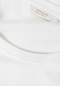 Λευκό βαμβακερό T-shirt με στρογγυλή λαιμόκοψη, ραμμένη γιακά και ορατή ετικέτα με κείμενο "TNTUUM COMMITMENT" σε ανοιχτό καφέ χρώμα.