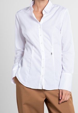 Chemise blanche cintrée à manches longues avec col et petite fleur noire brodée à la taille, portée avec un pantalon marron.
