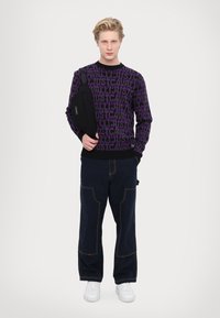 Purple knit sweater with bold black lettering, paired with navy blue wide-leg pants featuring contrasting stitching, and white sneakers. Black shoulder bag.