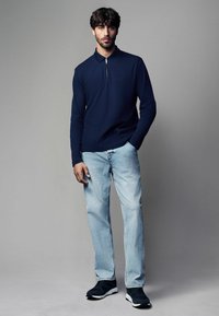 Polo de culoare bleumarin cu mânecă lungă și guler cu fermoar, dintr-un material texturat, asortat cu blugi drepți de un bleumarin deschis și pantofi sport negri.