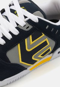 Navy blue sneaker with grey and yellow accents. Features a textured upper, white laces, and a patterned side logo. Rubber sole visible.