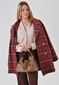 Roter karierter Wollmantel mit zwei Knöpfen, beiges Bluse und braune Ledertasche mit Kettendetails. Das Modell hält die Tasche und zeigt ihre Textur.
