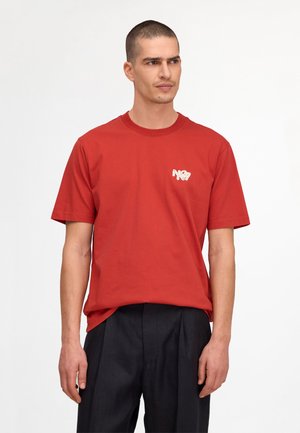 Mand med kort hår iført en rød t-shirt med en lille grafik på brystet og sorte bukser, stående mod en ensfarvet hvid baggrund.