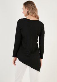 Top lungo a maniche nere a coste con scollatura dritta, caratterizzato da un orlo asimmetrico e tessuto testurizzato. Indossato con pantaloni bianchi.