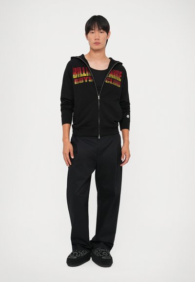 Crna zip-up dukserica s višebojnom "Billionaire Boys Club" logotipom, u kombinaciji s labavim crnim hlačama i tamnim ukrašenim papučama.