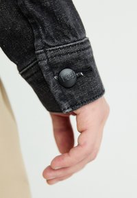 Black denim shirt cuff with a black plastic button featuring "Lee" logo. Visible stitching and textured surface, partially covering a hand.