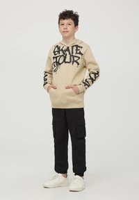 Felpa beige con stampa graffiti nera "SKATE TOUR", tasca a marsupio frontale e pantaloni cargo neri. Sneakers bianche completano l'outfit.
