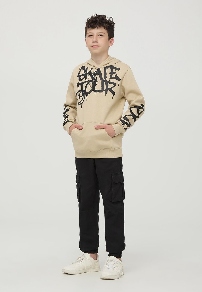 Felpa beige con stampa graffiti nera "SKATE TOUR", tasca a marsupio frontale e pantaloni cargo neri. Sneakers bianche completano l'outfit.