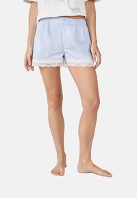 Hellblaue gestreifte Schlafshorts mit weißer Spitzenborte, getragen von einer stehenden Person, kombiniert mit einem weißen Cropped-Top.