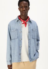 Ανοιχτό μπλε τζιν πουκάμισο με κουμπιά μπροστά, δύο τσέπες στο στήθος και μακριά μανίκια. Φορεμένο πάνω από γκρι πουλόβερ με λαιμόκοψη και αξεσουάρ με κόκκινη μαντήλα.