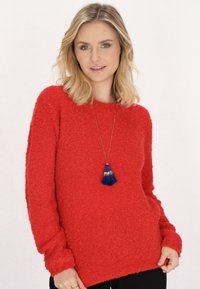 Roter strukturierter Pullover mit lockerer Passform und langen Ärmeln, ausgestattet mit einem runden Ausschnitt und einer blauen Quasten-Halskette für zusätzlichen Detailreichtum.