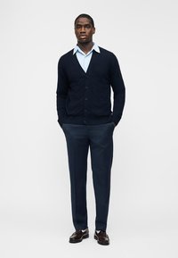 Férfi halványkék ingben, sötétkék kardigánnal, kék nadrágban és barna elegáns cipőben, zsebébe dugott kézzel áll.