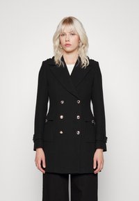 Manteau noir croisé avec de grands boutons en argent, poches latérales et détails aux épaules. Fabriqué en tissu texturé, coupe ajustée.