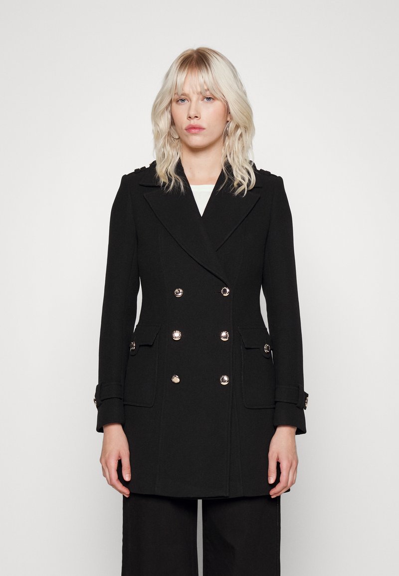 Manteau noir croisé avec de grands boutons en argent, poches latérales et détails aux épaules. Fabriqué en tissu texturé, coupe ajustée.