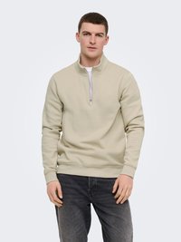 Jeune homme portant un sweat-shirt à fermeture éclair quart beige clair et un jean gris foncé, debout contre un fond uni clair.