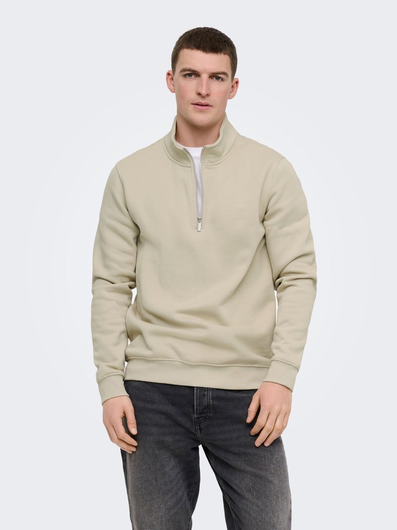 Jeune homme portant un sweat-shirt à fermeture éclair quart beige clair et un jean gris foncé, debout contre un fond uni clair.