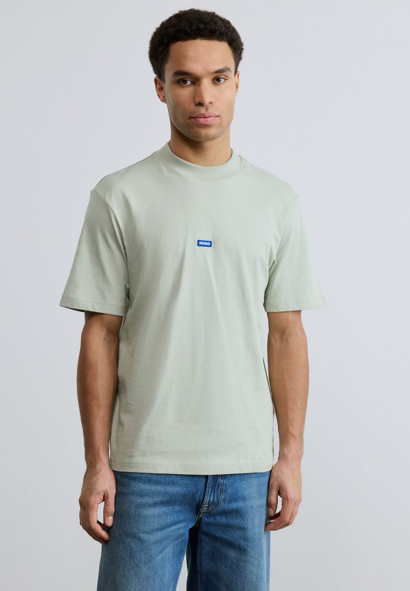 Νεαρός άνδρας με κοντά σγουρά μαλλιά φορώντας ανοιχτοπράσινη μπλούζα με λογότυπο HUGO και μπλε τζιν, στέκεται μπροστά σε απλό ανοιχτό γκρι φόντο.