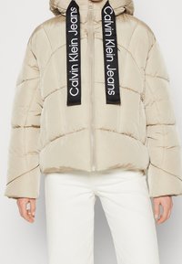 Beige puff jakke med en cropped design, der har diagonal syning og sorte logo snore. Metallisk lynlås og brede ærmer som detalje.