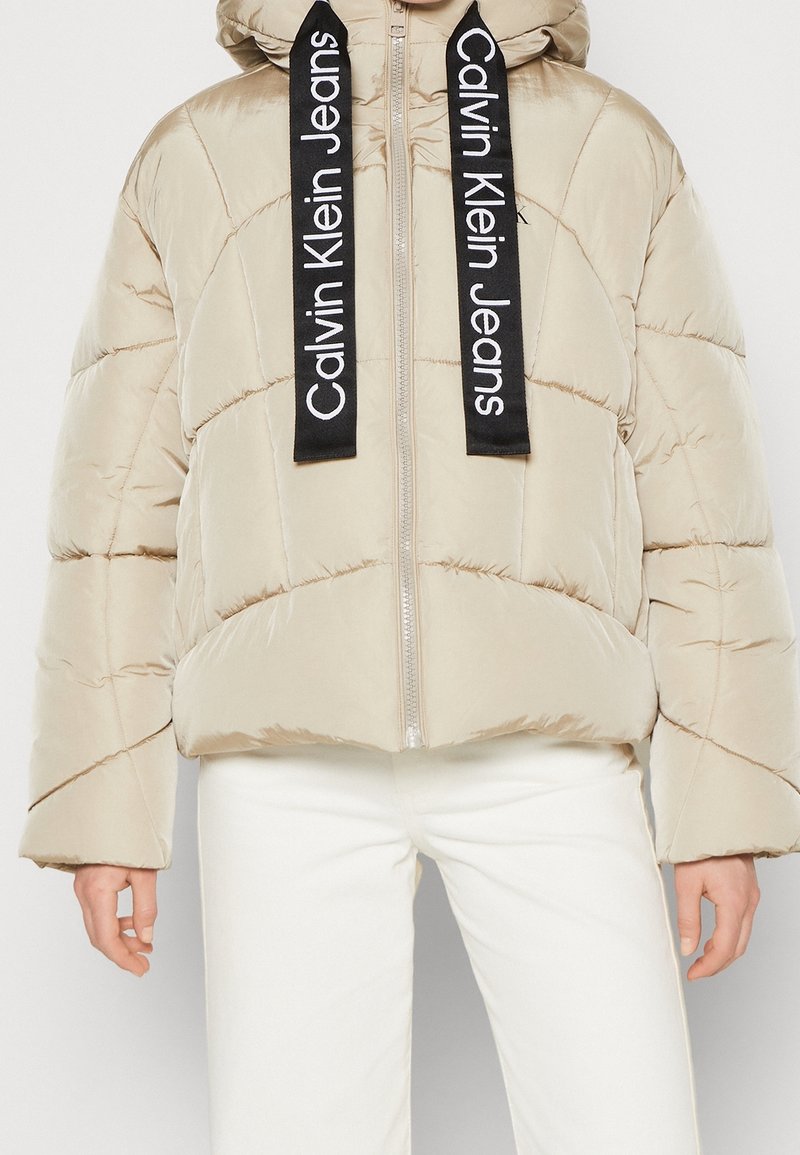 Beige puff jakke med en cropped design, der har diagonal syning og sorte logo snore. Metallisk lynlås og brede ærmer som detalje.