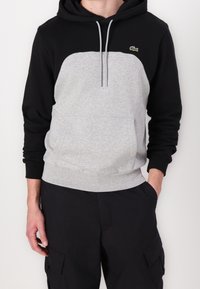 Sudadera con capucha de parte superior negra y cuerpo gris, que cuenta con un bolsillo frontal, cordón y pequeño logo verde en el pecho.