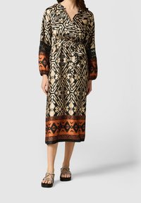 Långärmad klänning med V-ringning, prydd med ett geometriskt mönster i kräm, svart och rostfärg. Åtsittande midja med knytband och längd till anklarna.