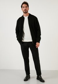 Fekete bomberdzseki egy fehér bordázott pulóver felett, slim-fit fekete nadrággal és fekete bokacsizmával párosítva, semleges háttérrel.