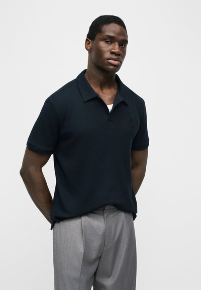 Homme portant un polo manches courtes bleu marine foncé et un pantalon gris clair, debout les mains derrière le dos sur un fond uni.