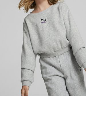 Młoda osoba ubrana w jasnoszarą bluzę Puma i dopasowane spodnie dresowe na jednolitym tle.