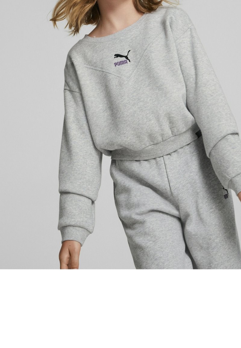 Jeune personne portant un sweat-shirt Puma gris clair et un pantalon de survêtement assorti sur un fond uni.