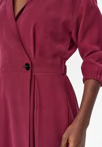 Zbliżenie bordowej sukienki z dekoltem w serek, pojedynczym czarnym guzikiem w talii, plisami poniżej talii oraz elastycznym rękawem 3/4 na ciemnoskórej ręce.