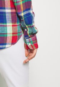 Personne portant une chemise à carreaux multicolores à manches longues avec des poignets boutonnés et une jupe blanche, main reposant le long du corps.
