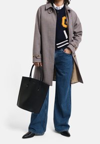 Brunternet frakk med knapper, marineblå sweater med guldskrift, brede blå jeans, sort tote bag og sorte spidse sko.