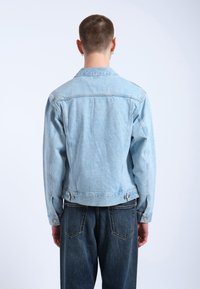 Chaqueta de denim azul claro con cuello, costuras visibles y botones laterales. Llevada sobre jeans azul oscuro, mostrando un diseño ajustado.