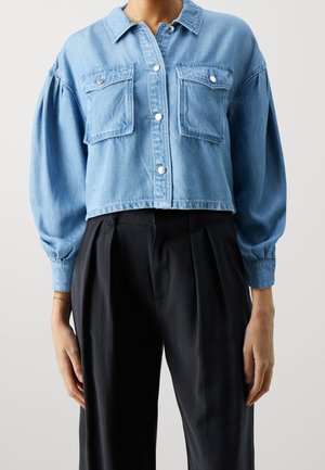 Vrouw met een cropped lichtblauwe spijkerjack met klepzakken aan de voorkant en zwarte hooggetailleerde geplooide broek, staand tegen een effen achtergrond.