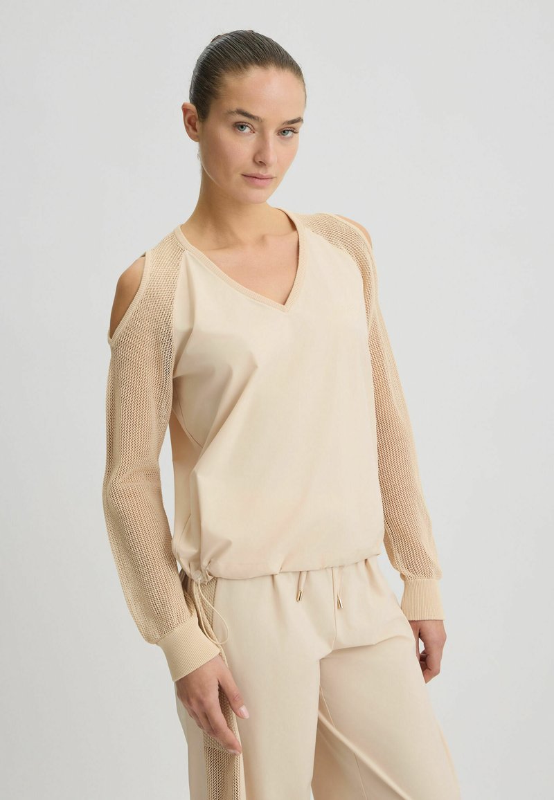 Beige lange mouw top met een V-hals, voorzien van mesh mouwen en een geplooide taille. Zachte stof met textuurelementen.