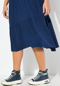 Abito blu navy con un design a strati e tessuto fluido, abbinato a sneakers alte blu navy con lacci bianchi e suola color tan.