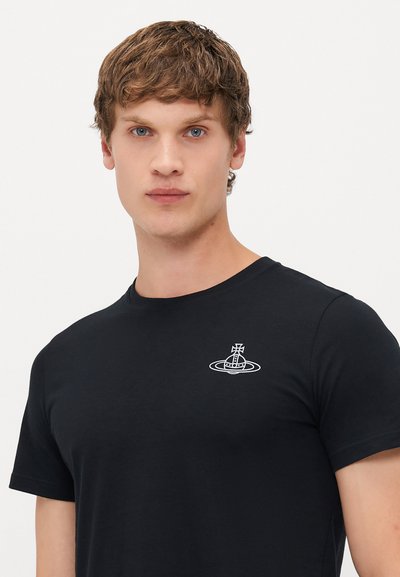 Young man with short brown hair wearing a black t-shirt featuring a small white orb and cross emblem on the left chest.