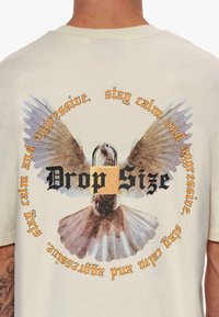 T-shirt beige clair avec un motif central représentant un oiseau aux ailes déployées, entouré d'un texte stylisé en noir et orange.