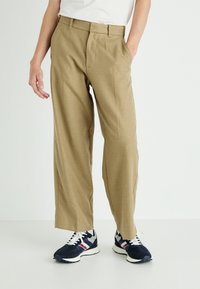 Beige, wide-leg trousers made of textured fabric, featuring front pleats and a standard waistband, paired with black sneakers with white accents.