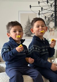 Kaksi lasta, jotka käyttävät tummansinisiä neuleita, joissa on kuvioituja yksityiskohtia, pitävät ruokaa sylissään istuen vaalean värisessä, teksturoidussa ympäristössä pehmeässä sisävalaistuksessa.