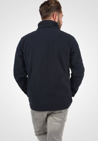 Chaqueta de forro polar negra con un cuello alto. Tiene una superficie texturizada y un ajuste relajado, combinada con jeans gris claro en un entorno informal.