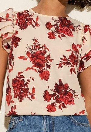Kvinde iført cremefarvet bluse med rødt blomsterprint og flæset kapærmer, kombineret med blå cowboybukser.