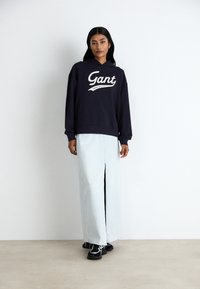 Navy hooded sweatshirt with white "Gant" logo, paired with a light blue skirt featuring a front slit, complemented by black sneakers.