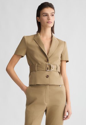 Giacca beige corta con maniche corte, vita cintata con fibbia metallica e colletto a punta. Tessuto liscio e dettagli con bottoni.