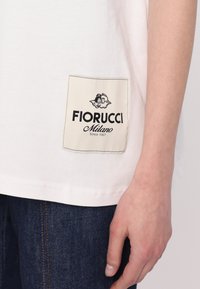 Fehér póló, amelyen egy krém színű címke található fekete logóval, "Fiorucci Milano" felirattal a bal alsó részen, sötét farmernadrággal párosítva.